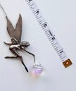 A silver fairy pendant holding a faceted iridescent crystal ball, shown next to a white measuring tape on a white background.