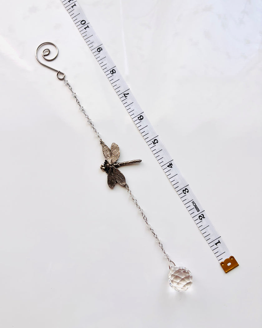 A silver-toned suncatcher featuring a dragonfly charm, a spiral ring at one end, and a faceted clear crystal bead at the other, shown on a white background next to a measuring tape for scale.