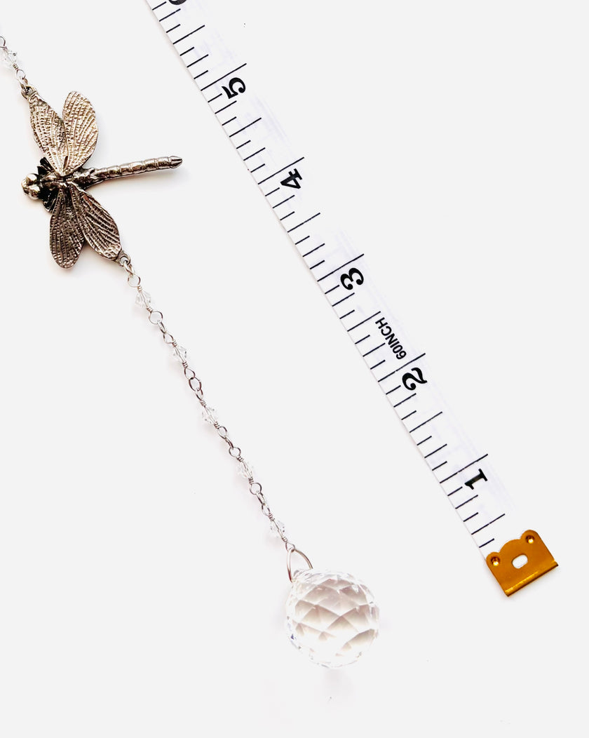 A silver-toned dragonfly suncatcher with a clear faceted crystal ball charm, displayed next to a measuring tape to show scale on a white background.