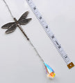 A silver dragonfly pendant with a teardrop-shaped iridescent crystal drop, shown next to a white measuring tape on a light gray background.
