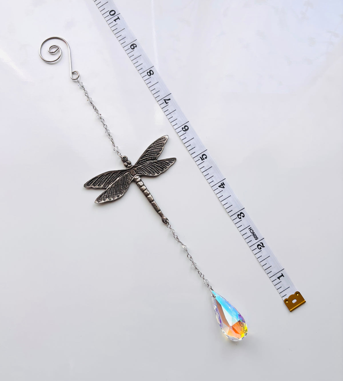 A silver dragonfly suncatcher with a teardrop-shaped iridescent crystal pendant, shown next to a white measuring tape on a white background.
