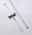 A silver dragonfly suncatcher with a teardrop-shaped iridescent crystal pendant, shown next to a white measuring tape on a white background.