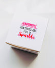 A white square gift box with a printed warning label that reads 'WARNING! CONTENTS ARE FULL OF Sparkle' in pink and black text, photographed on a clean white surface.