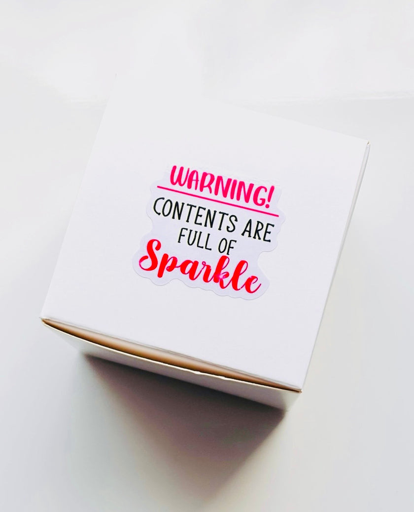 A white square gift box with a printed warning label that reads 'WARNING! CONTENTS ARE FULL OF Sparkle' in pink and black text, photographed on a clean white surface.