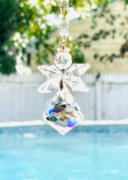 A clear crystal angel suncatcher with a faceted diamond-shaped body and wings, hanging in front of a window with a swimming pool and green trees in the background.