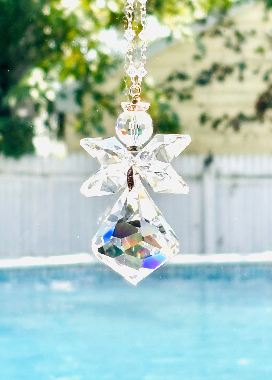 A clear crystal angel suncatcher with a faceted diamond-shaped body and wings, hanging in front of a window with a swimming pool and green trees in the background.