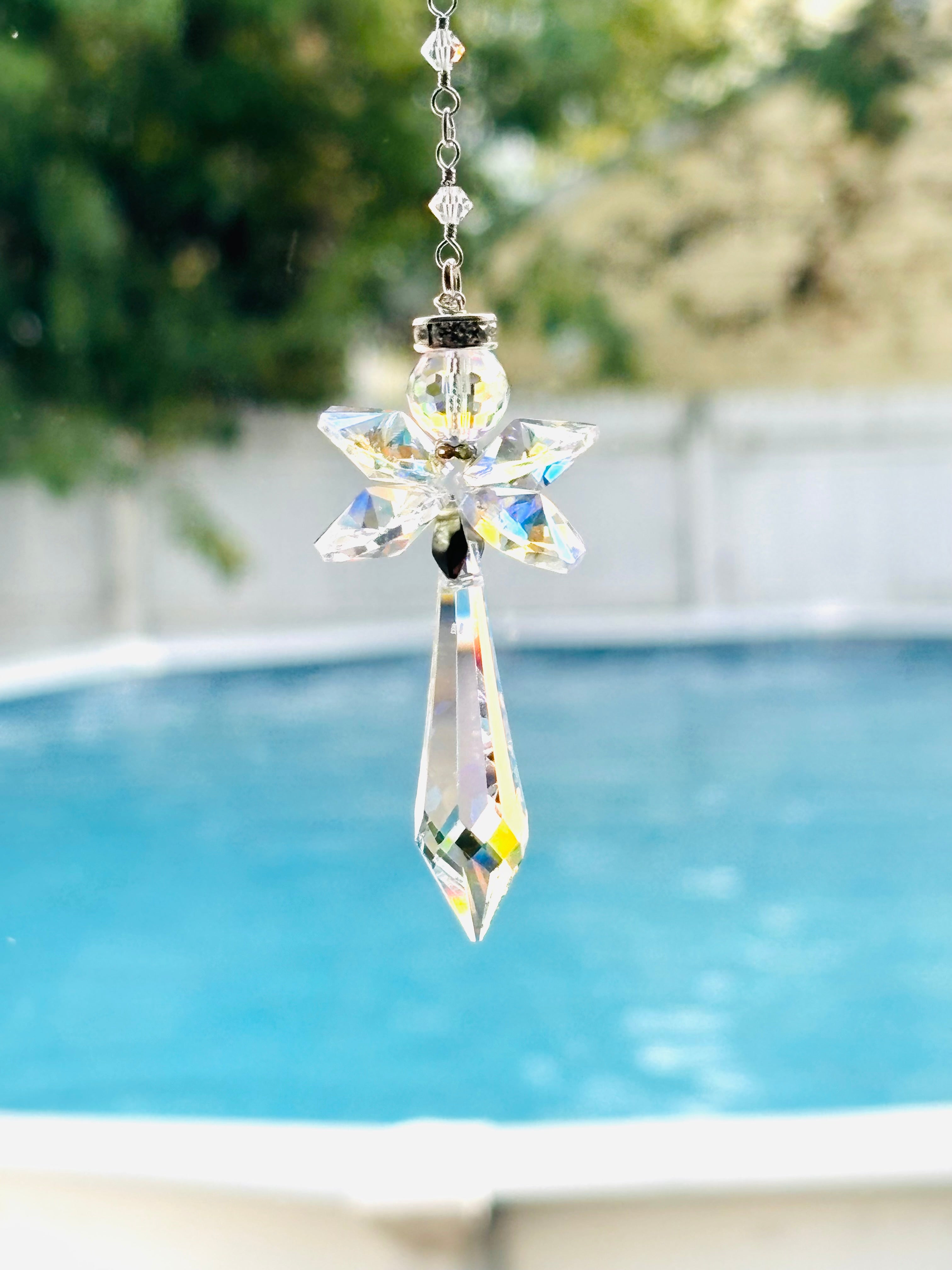 A clear crystal angel suncatcher with a pointed bottom and hanging chain, shown outdoors in front of a swimming pool with a blurred background.