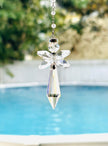 A clear crystal angel suncatcher with a pointed bottom and a silver chain, shown hanging outdoors in front of a blurred swimming pool and greenery.