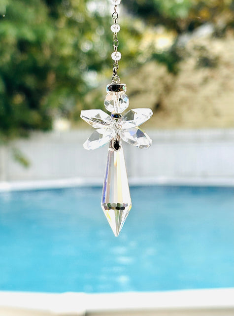 A clear crystal angel suncatcher with a pointed bottom and a silver chain, shown hanging outdoors in front of a blurred swimming pool and greenery.