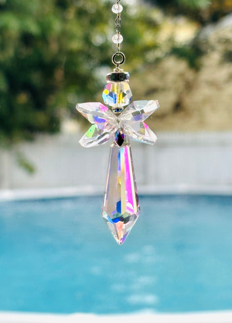 An iridescent crystal angel suncatcher hanging outdoors in front of a blurred swimming pool and greenery background.