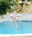An Aurora Borealis faceted butterfly suncatcher with an iridescent rainbow finish hangs outdoors in front of a blurred swimming pool and greenery.