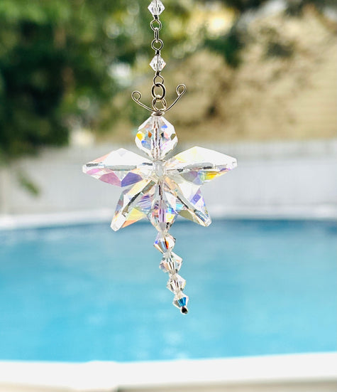 An Aurora Borealis faceted butterfly suncatcher with an iridescent rainbow finish hangs outdoors in front of a blurred swimming pool and greenery.