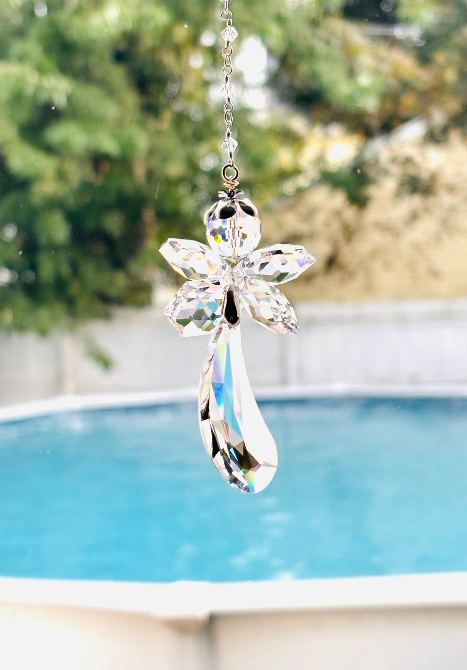 A clear faceted fairy angel suncatcher with a long iridescent wing, hanging in front of a blurred outdoor background featuring a swimming pool and green foliage.