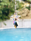 A blue fairy crystal suncatcher with a star-shaped top and a leaf-shaped bottom, made of iridescent clear glass, suspended in front of a blurred outdoor background with a swimming pool.