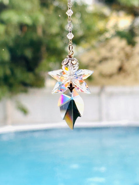 A blue fairy crystal suncatcher with a star-shaped top and a leaf-shaped bottom, made of iridescent clear glass, suspended in front of a blurred outdoor background with a swimming pool.