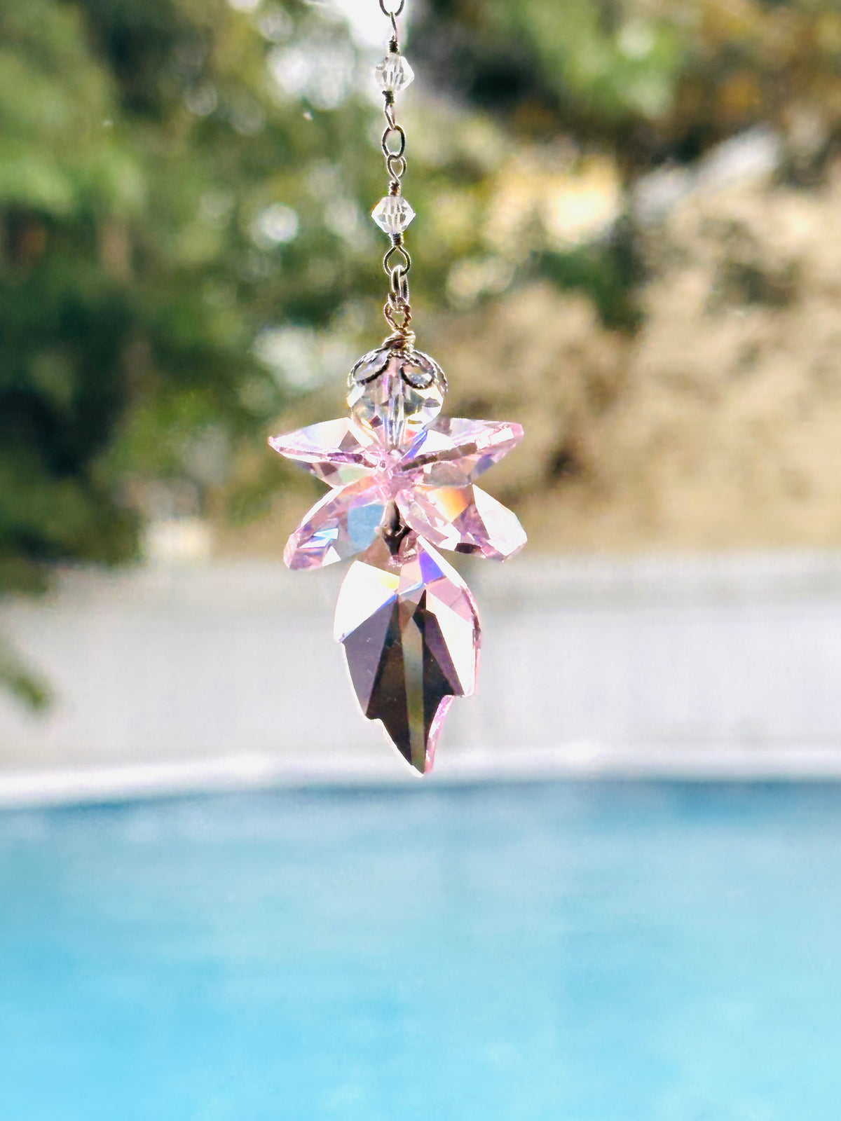 A pink faceted crystal fairy suncatcher with a floral starburst design hangs from a silver chain in front of a blurred outdoor background featuring a swimming pool and greenery.
