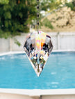 A large faceted clear crystal suncatcher hanging from a metal chain in front of a swimming pool, showing rainbow light reflections.