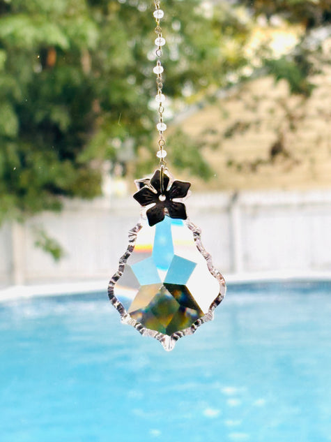 A hanging suncatcher with a faceted clear crystal body, a silver flower-shaped top, and a silver-toned chain, photographed outdoors with a blurred swimming pool and greenery in the background.