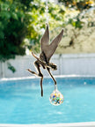 A metal fairy suncatcher with wings and a dangling faceted rainbow prism ball, photographed outdoors over a swimming pool with a white fence and greenery in the background.