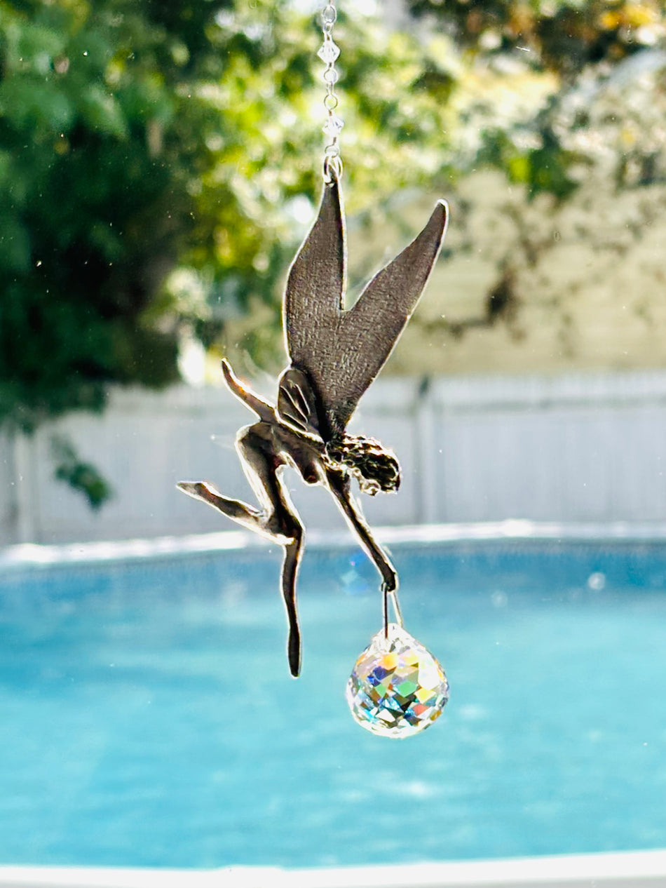 A metal fairy suncatcher with wings and a dangling faceted rainbow prism ball, photographed outdoors over a swimming pool with a white fence and greenery in the background.