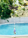 A dragonfly-shaped suncatcher with a dangling faceted crystal teardrop, photographed outdoors in front of a swimming pool and green trees.