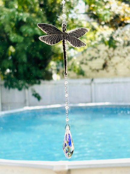 A dragonfly-shaped suncatcher with a dangling faceted crystal teardrop, photographed outdoors in front of a swimming pool and green trees.
