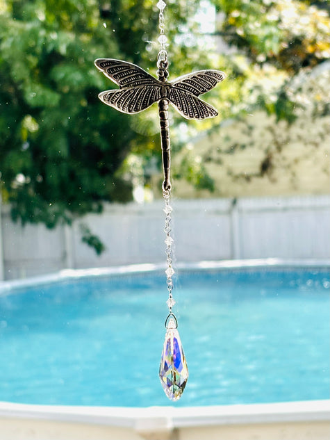 A dragonfly-shaped suncatcher with a dangling faceted crystal teardrop, photographed outdoors in front of a swimming pool and green trees.