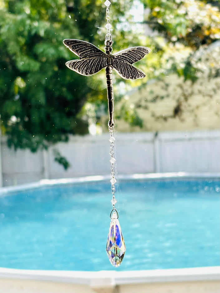 A dragonfly-shaped suncatcher with a dangling faceted crystal teardrop, photographed outdoors in front of a swimming pool and green trees.