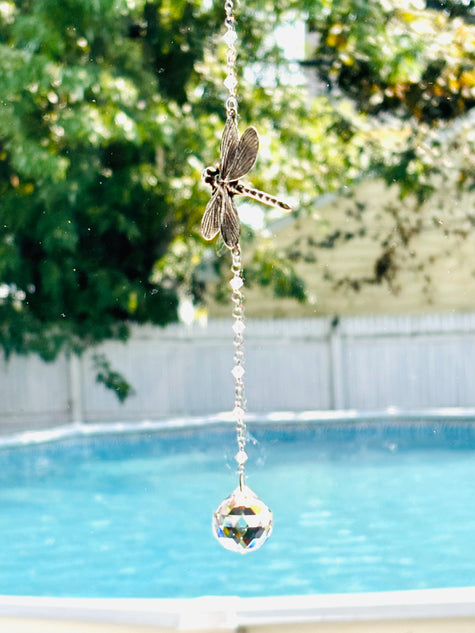 A hanging suncatcher with a metallic dragonfly and a faceted clear crystal ball, photographed outdoors in front of a swimming pool with blurred green trees and a fence in the background.