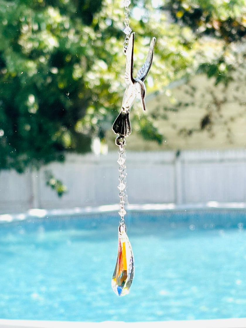 A hanging bird-shaped suncatcher with a colorful teardrop glass pendant, suspended in front of a blurred swimming pool and green trees.