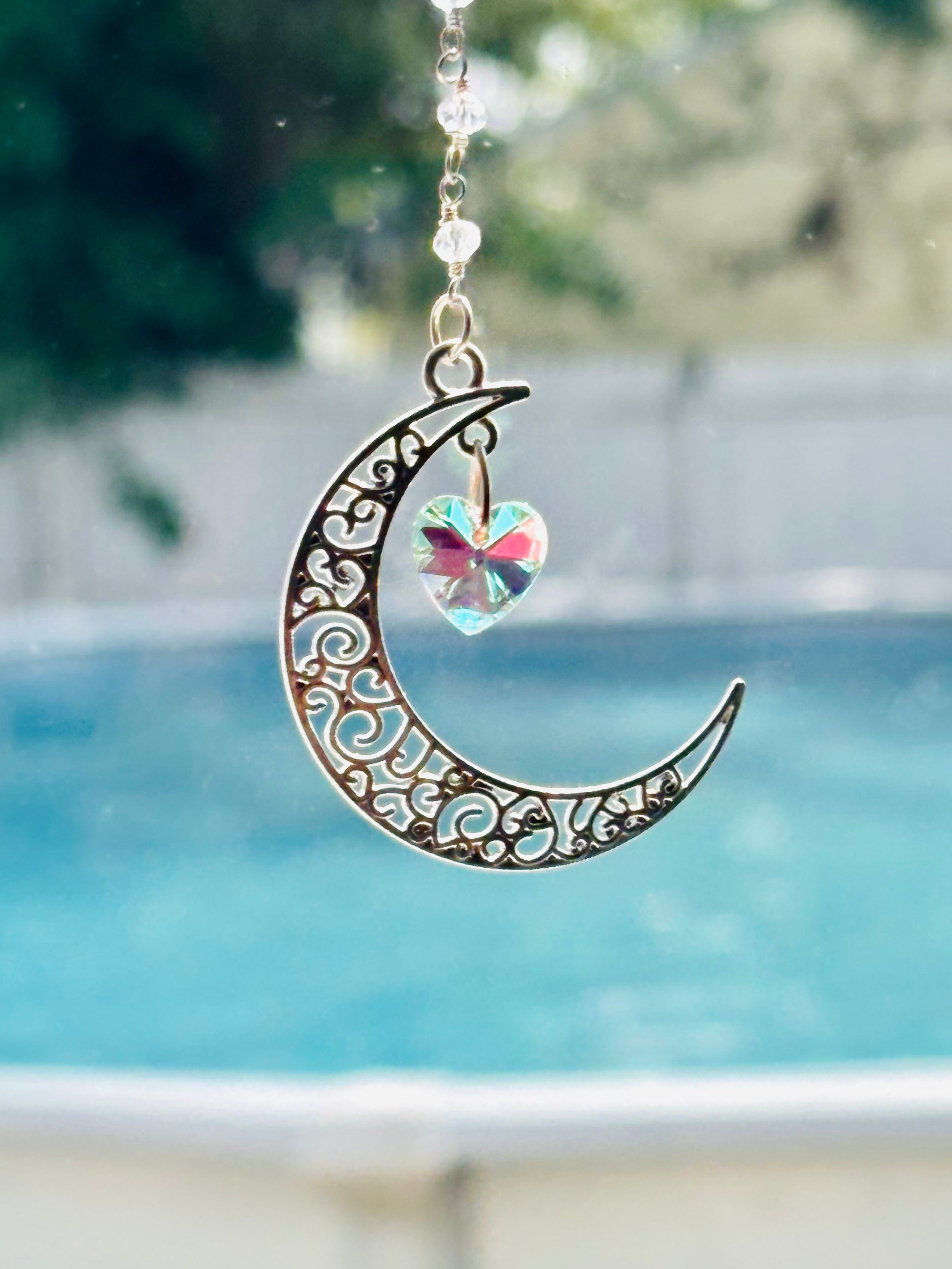 A crescent moon-shaped metal ornament with intricate scrollwork and a small iridescent heart-shaped crystal hanging from it, photographed outdoors in front of a blurred swimming pool and greenery.