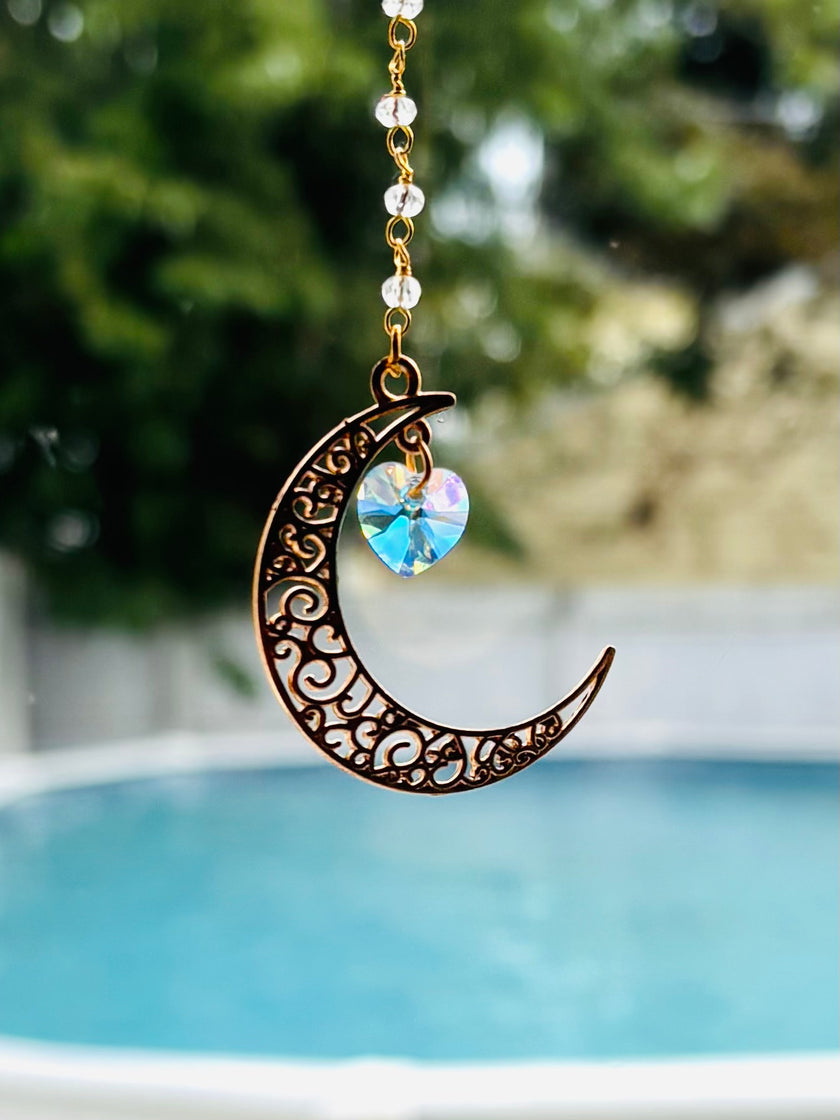 A gold-toned crescent moon suncatcher necklace with intricate scrollwork cutouts and a dangling iridescent heart-shaped crystal, shown outdoors with a blurred swimming pool and greenery in the background. 