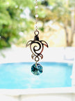 A metal turtle suncatcher ornament with a blue faceted crystal charm, hanging outdoors in front of a blurred swimming pool and green foliage.