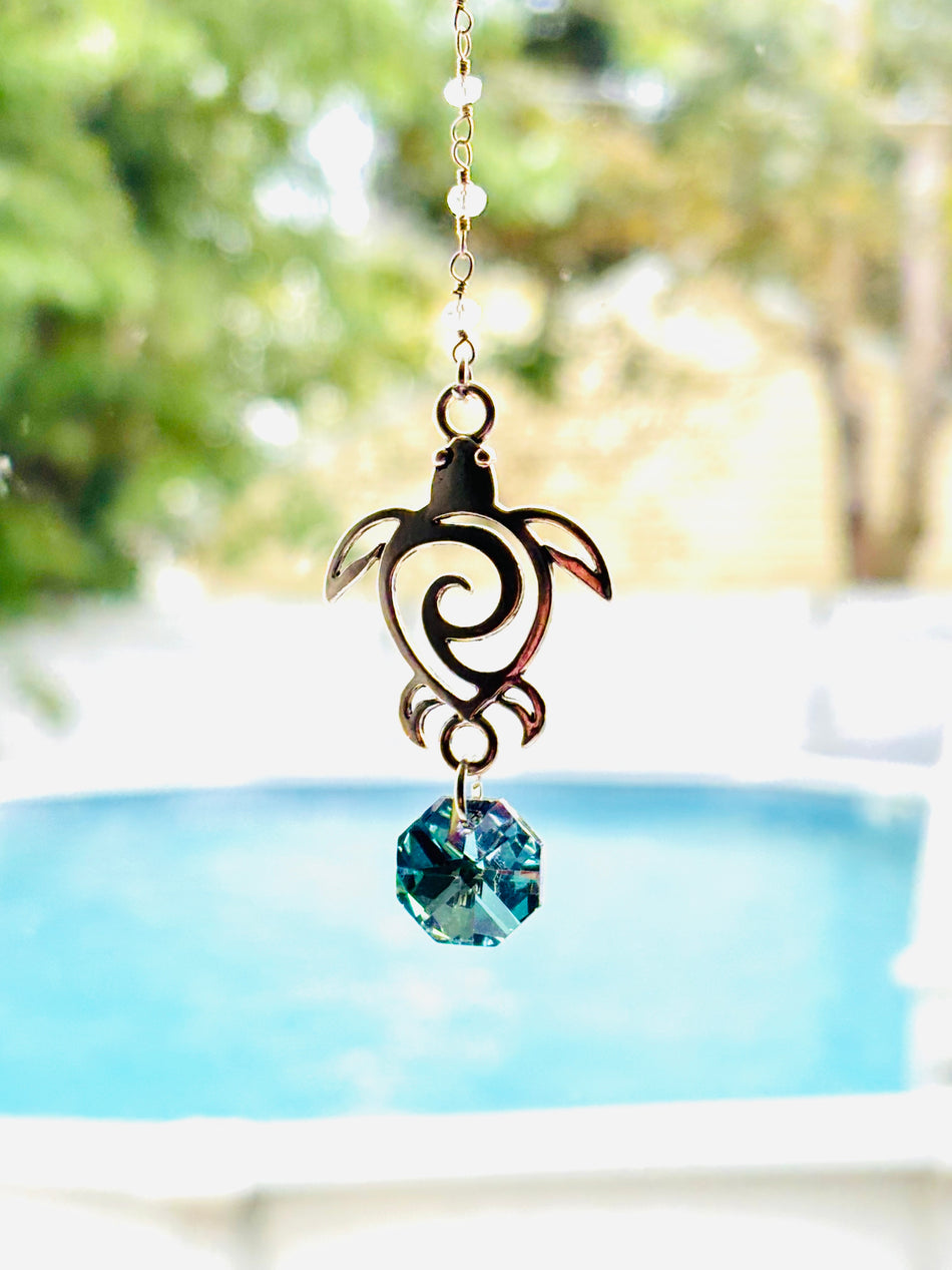 A metal turtle suncatcher ornament with a blue faceted crystal charm, hanging outdoors in front of a blurred swimming pool and green foliage.