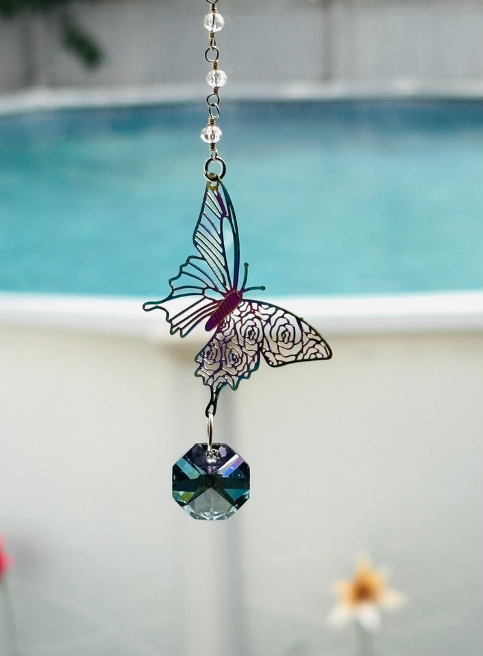 A hanging butterfly suncatcher with a faceted blue-green crystal pendant, shown outdoors in front of a blurred swimming pool.