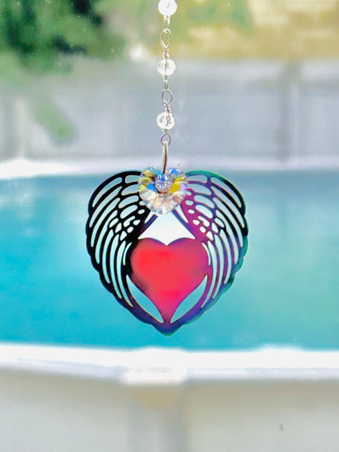 A heart-shaped metal suncatcher with black wing cutouts, a red heart in the center, and a small iridescent flower accent at the top, hanging in front of a blurred swimming pool background.