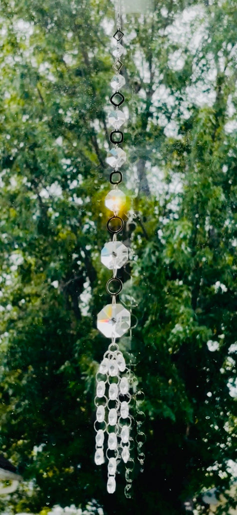 Handmade Neptune’s Octagon Prism crystal suncatcher hanging in a window against a background of lush green trees, capturing bright golden sunlight through its clear faceted glass prisms.