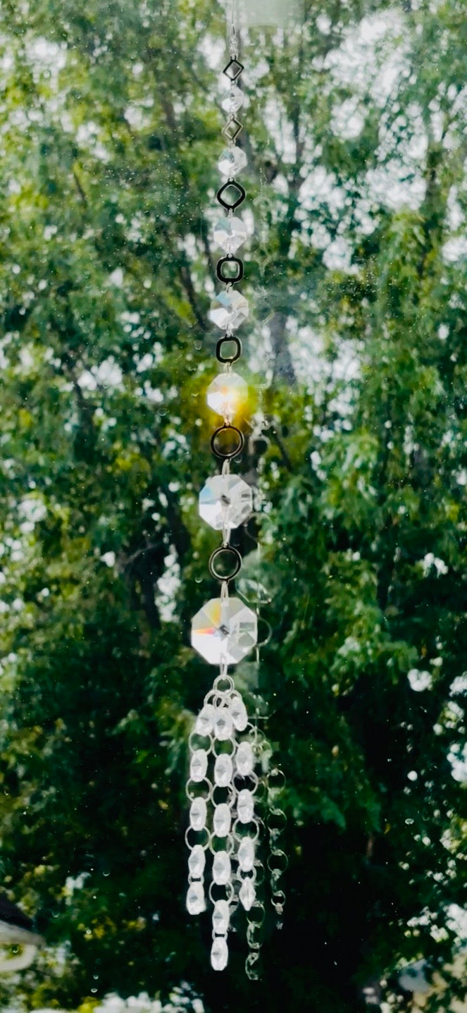 Handmade Neptune’s Octagon Prism crystal suncatcher hanging in a window against a background of lush green trees, capturing bright golden sunlight through its clear faceted glass prisms.