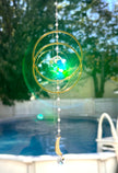 Handcrafted Serene Orbit crystal suncatcher casting a vibrant green lens flare and rainbow light reflection outdoors over a blue swimming pool background.