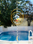 Handcrafted 'Serene Orbit' crystal suncatcher featuring interlocking golden geometric hoops and a central iridescent Swarovski hex-star prism hanging outdoors in a sunny backyard over a blue swimming pool.