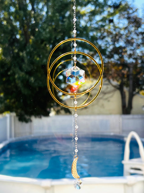 Handcrafted 'Serene Orbit' crystal suncatcher featuring interlocking golden geometric hoops and a central iridescent Swarovski hex-star prism hanging outdoors in a sunny backyard over a blue swimming pool.