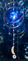 Handcrafted Serene Orbit crystal suncatcher casting a bright blue glow with a celestial, starry blue background accentuating the Swarovski prism and golden hoops.