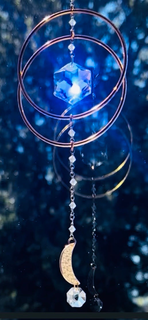 Handcrafted Serene Orbit crystal suncatcher casting a bright blue glow with a celestial, starry blue background accentuating the Swarovski prism and golden hoops.