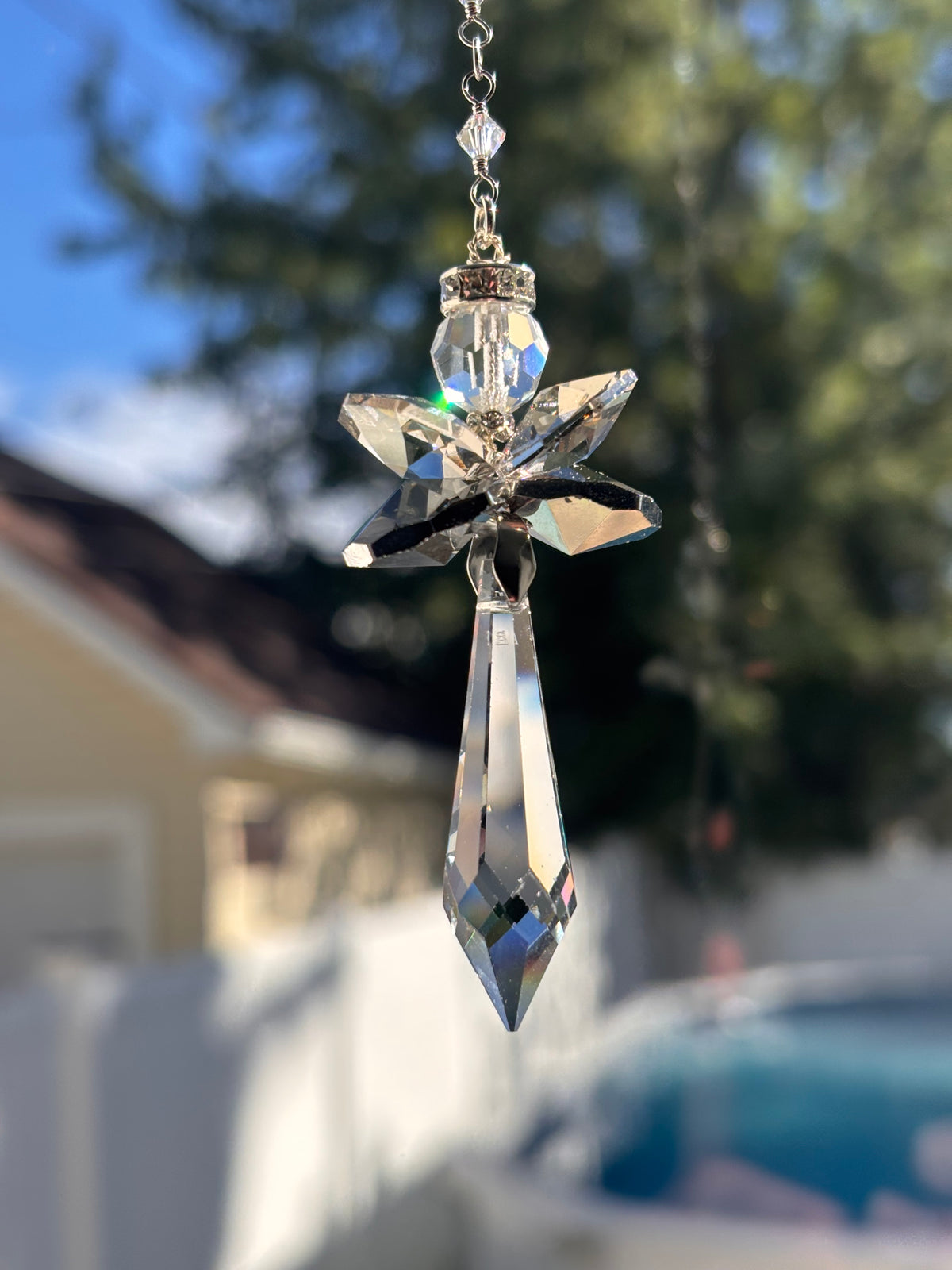 Handcrafted Hopea crystal angel suncatcher made with genuine Swarovski crystals featuring smoky metallic wings and a long clear faceted prism body hanging outdoors over a pool background.