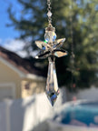 Handcrafted Hopea crystal angel suncatcher made with genuine Swarovski crystals featuring smoky metallic wings and a long clear faceted prism body hanging outdoors over a pool background.