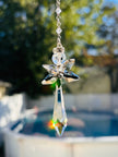 Handcrafted Hopea crystal angel suncatcher made with genuine Swarovski crystals featuring smoky metallic wings and a long clear faceted prism body hanging outdoors over a pool background.