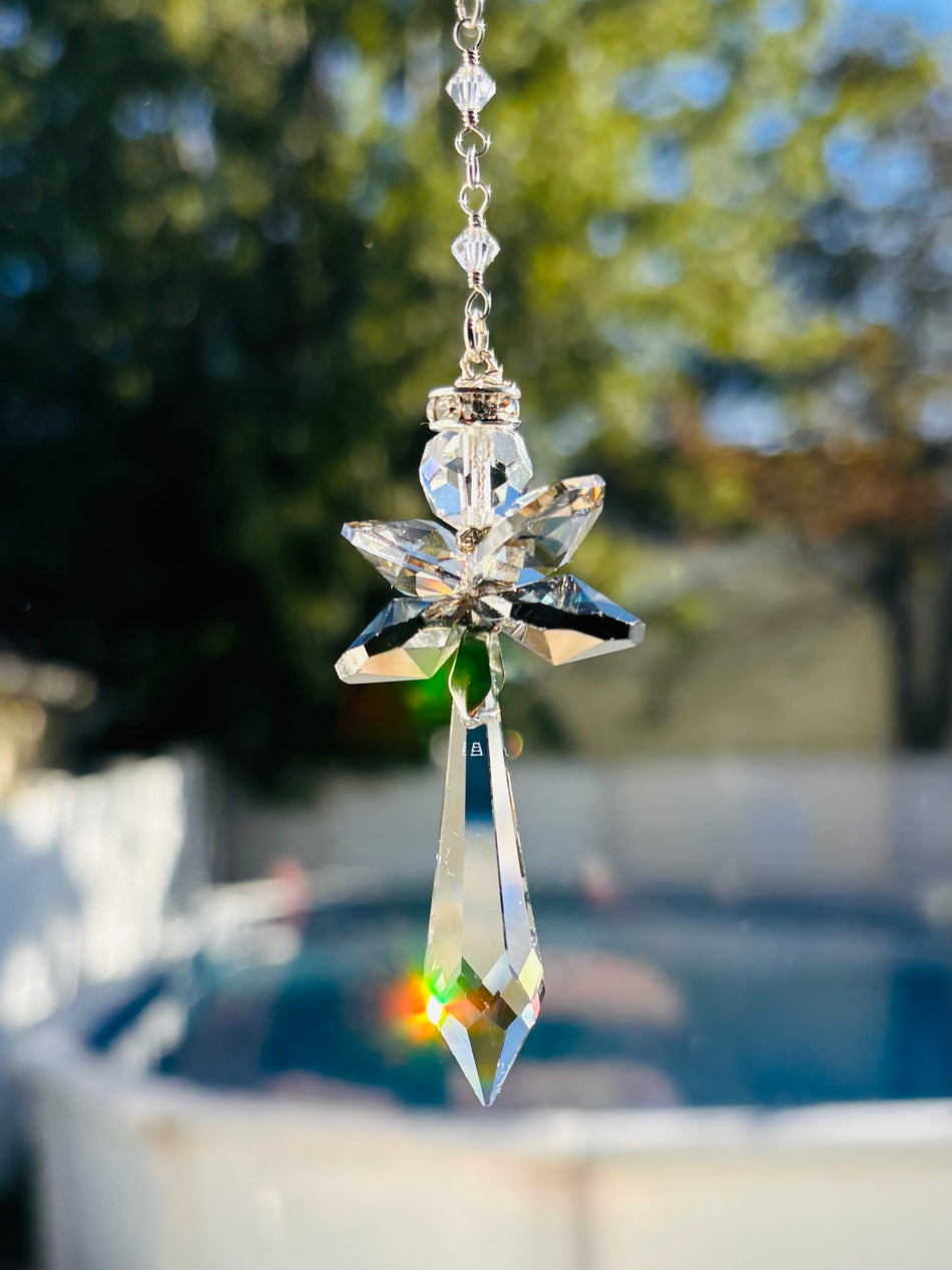 Handcrafted Hopea crystal angel suncatcher made with genuine Swarovski crystals featuring smoky metallic wings and a long clear faceted prism body hanging outdoors over a pool background.