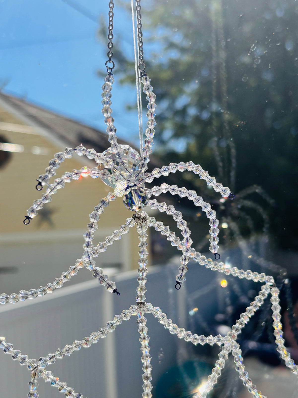 Macro close-up of a handmade crystal spider perched at the top of an intricate beaded spiderweb suncatcher. The daylight catches the genuine Swarovski and glass bicone crystals, throwing vibrant rainbow flares across the structured web lines.