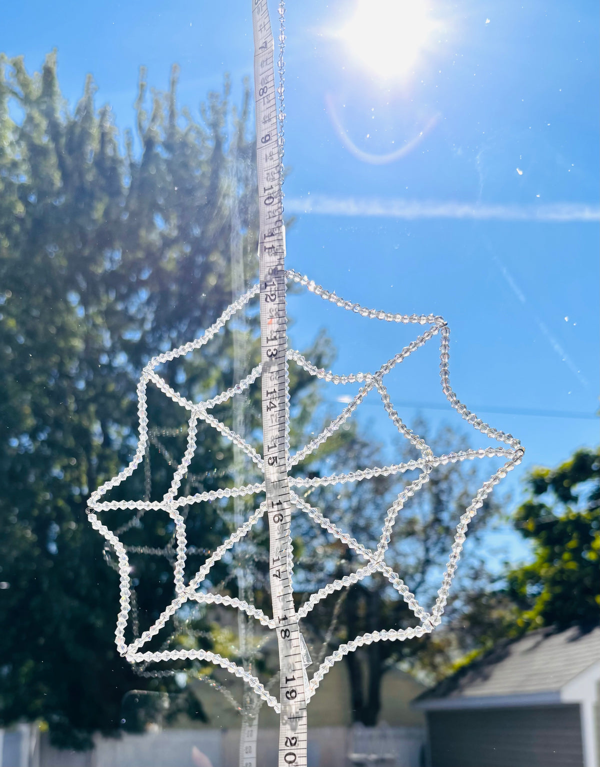 A handmade crystal spiderweb suncatcher being measured vertically with a yellow tape measure outdoors. The tape measure shows the precise scale, width, and hanging dimensions of the full 448-bead crystal web structure against a background of trees.