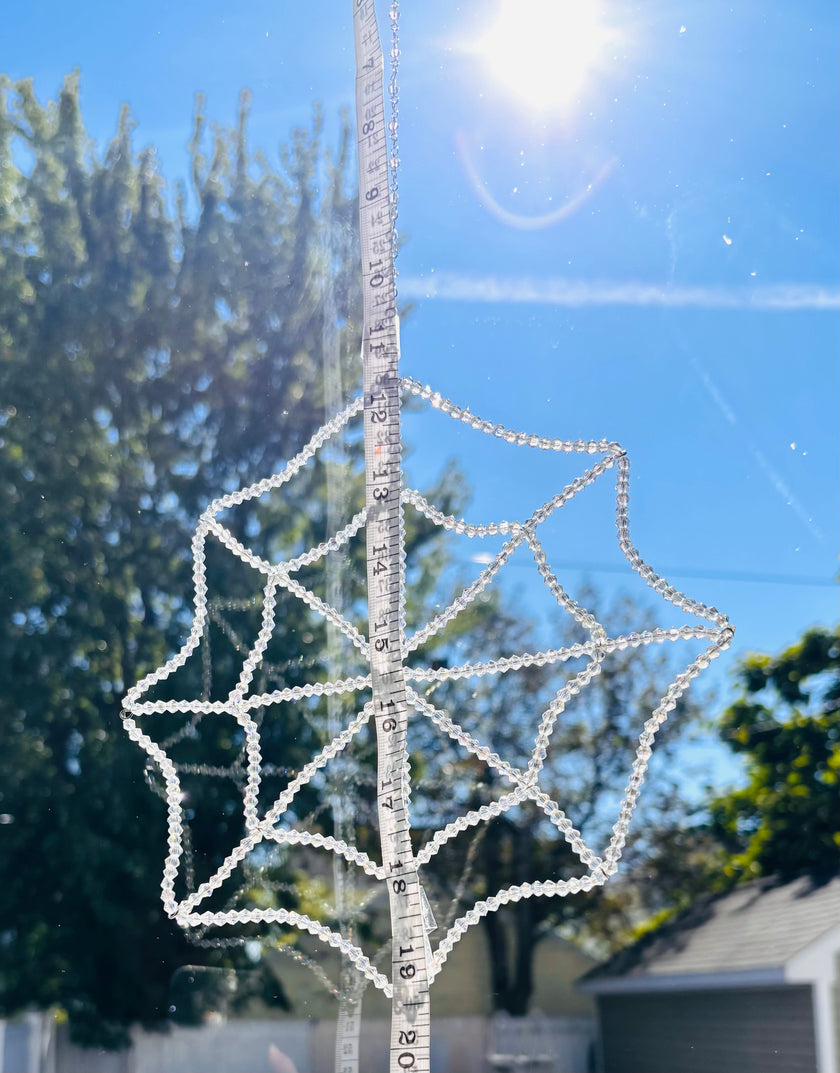 A handmade crystal spiderweb suncatcher being measured vertically with a yellow tape measure outdoors. The tape measure shows the precise scale, width, and hanging dimensions of the full 448-bead crystal web structure against a background of trees.